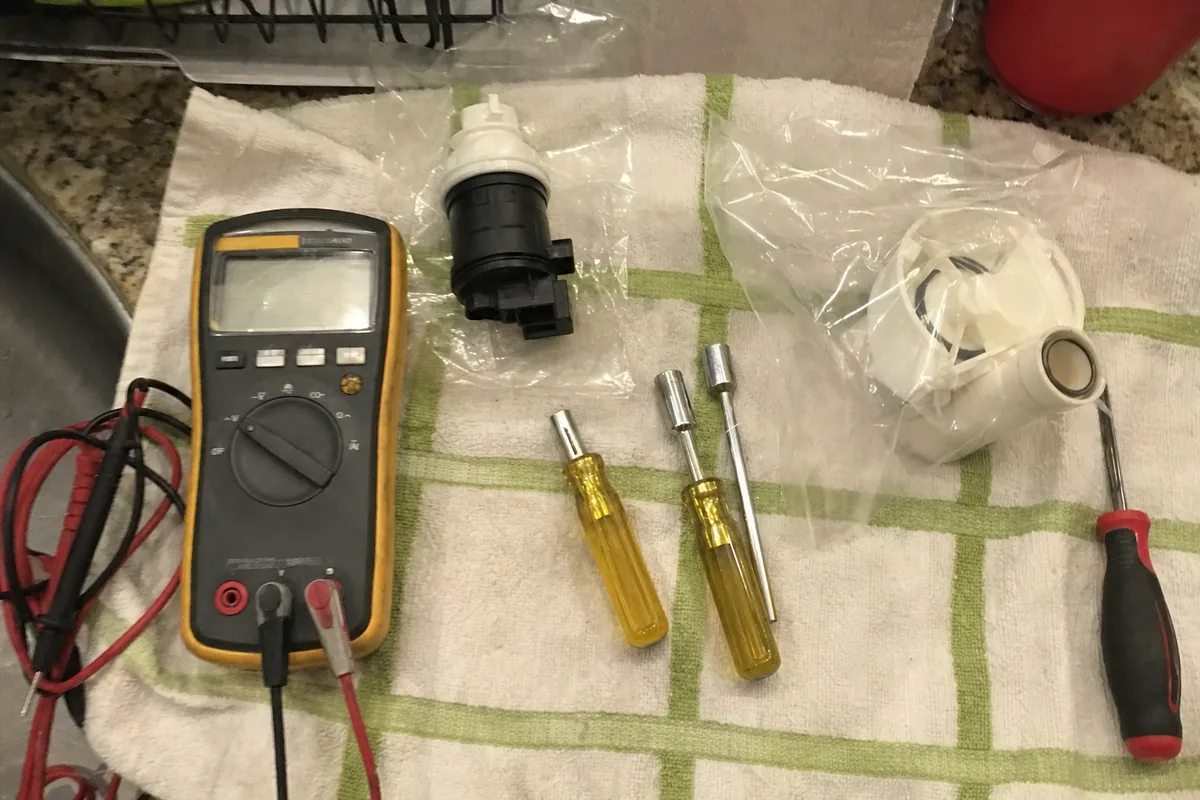 A bagged replacement part, a multimeter with red and black leads, and two nut drivers laid out on a towel