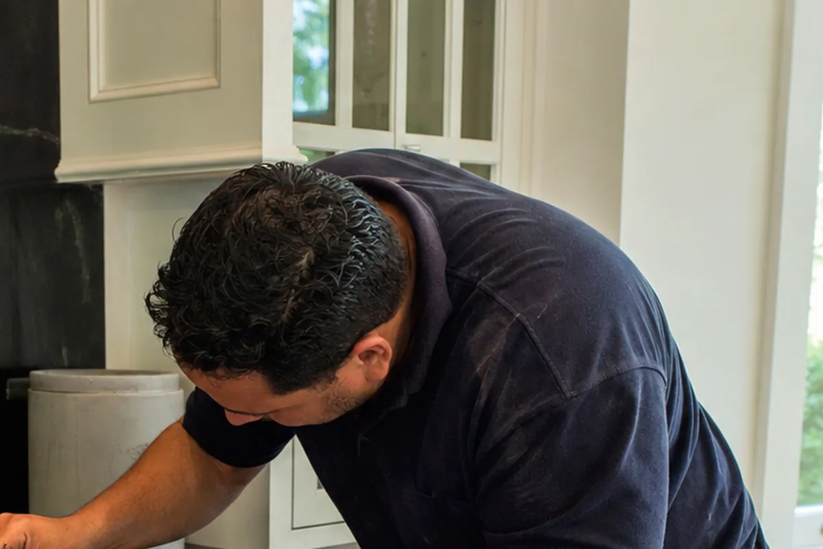 A technician bent over the open burner deck of a stainless range against a dark stone backsplash, one burner well uncovered