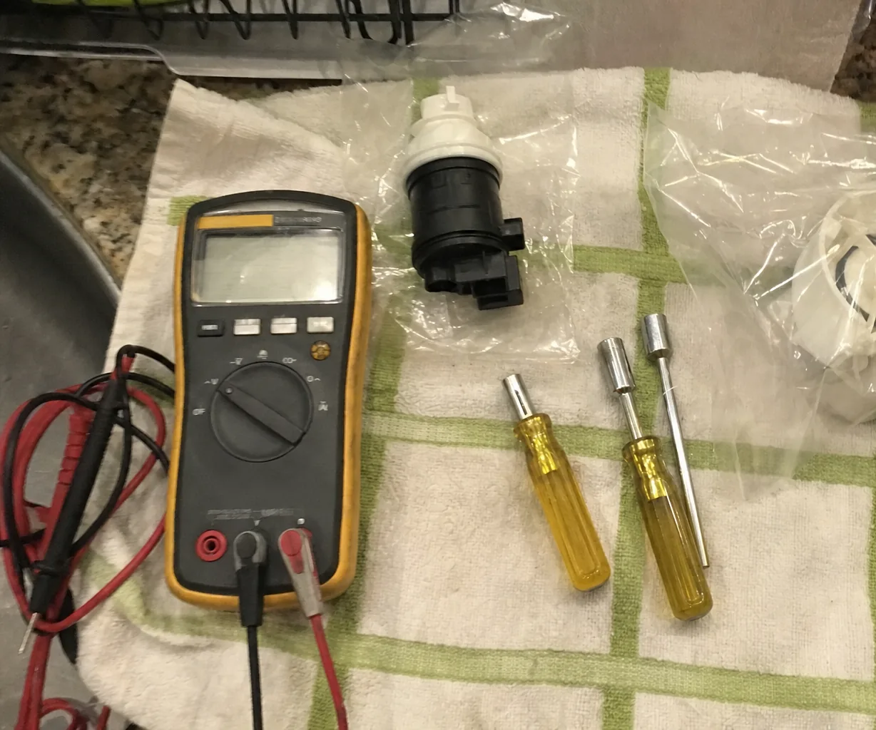 A yellow-cased multimeter with red and black test leads lying on a green-checked kitchen towel beside three yellow-handled nut drivers and a black and white part still in its plastic bag